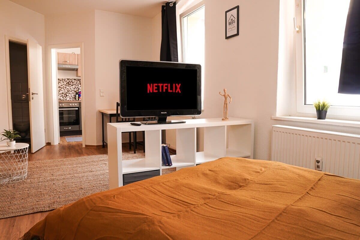 A bedroom with a bed, a TV displaying 'NETFLIX', a window, and a kitchenette in the background.