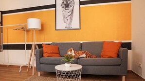 Un salon avec un canapé gris, des coussins orange, une lampe blanche et un tableau représentant un tigre, accroché au mur.