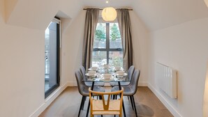 Een eetruimte met een glazen tafel, grijze stoelen, een hanglamp en uitzicht door een raam met gordijnen.