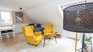 Una habitación con dos sillones amarillos, una mesita, un sofá gris y un escritorio blanco con una silla. Se ve una ventana con vistas al exterior, una lámpara con pantalla estampada y una planta sobre una mesa.