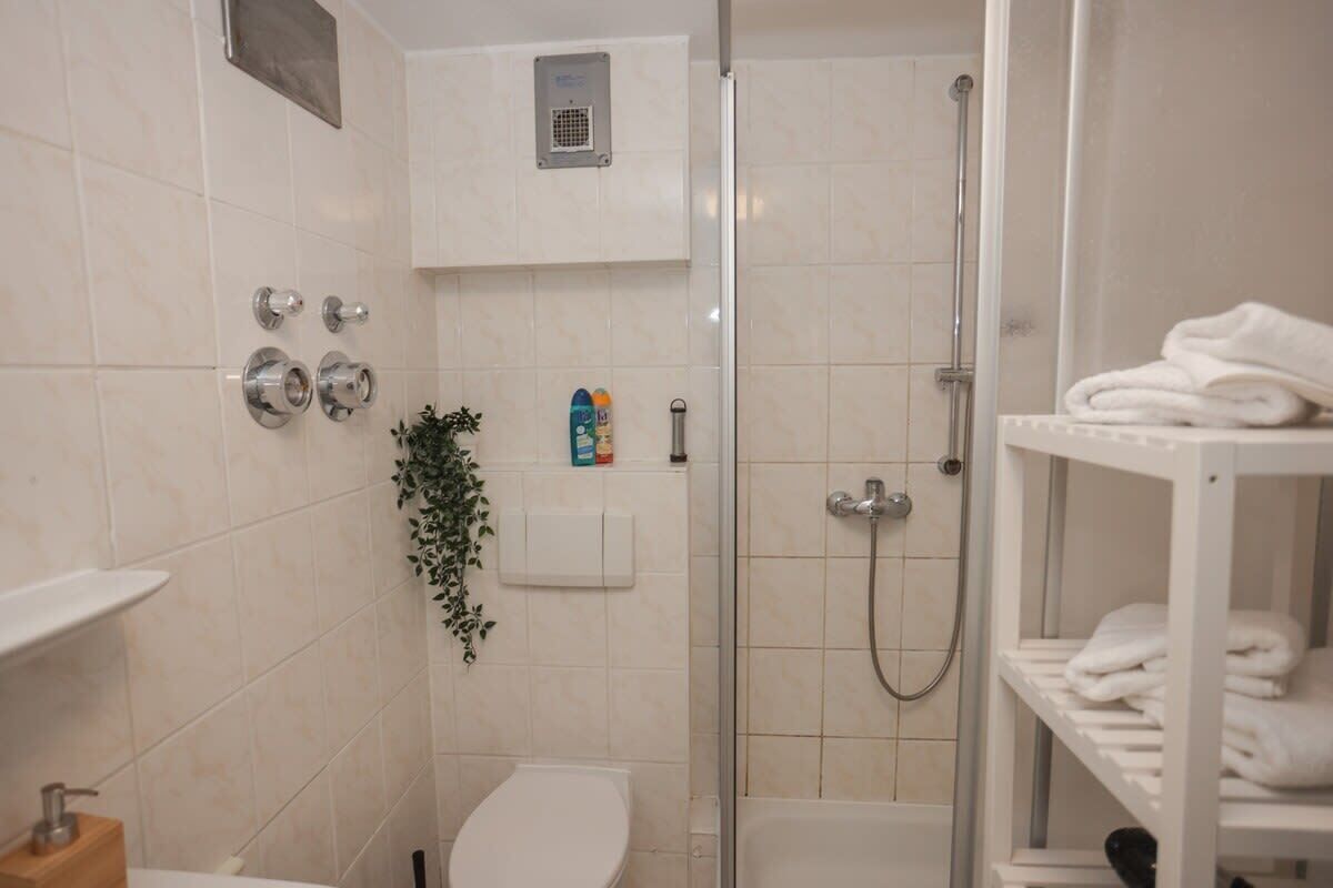 A small, modern bathroom with a toilet, a shower, and a sink. There is a shelf with towels and toiletries.
