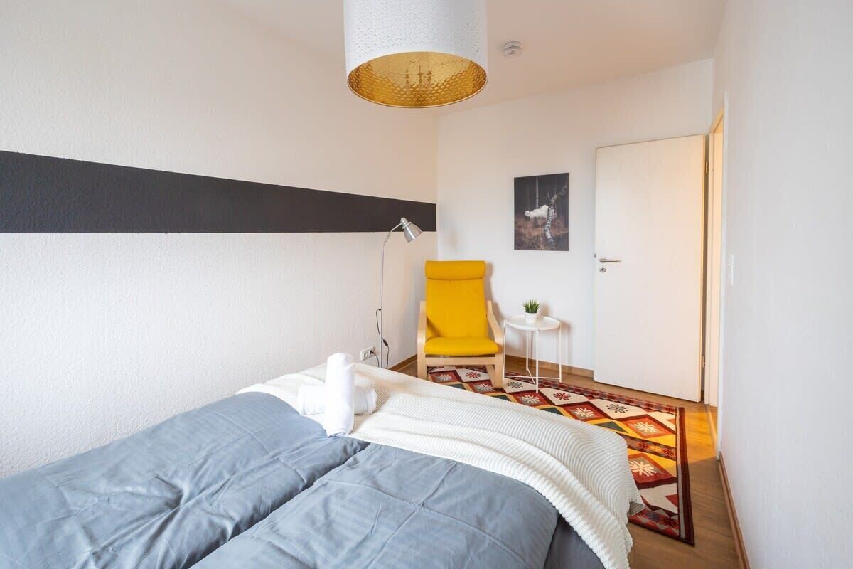 A bedroom with a bed, a chair, a small table, a lamp, and a door.