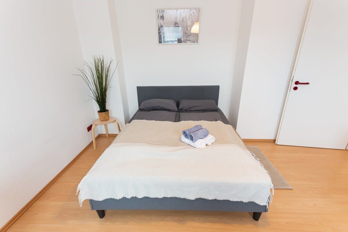 Una cama bien hecha con colcha blanca y gris, una pequeña planta en maceta y un cuadro en la pared.