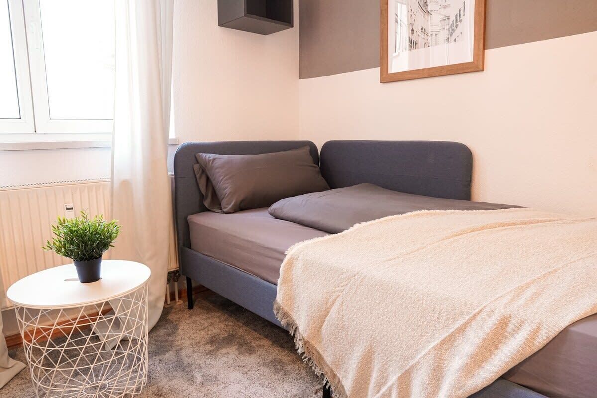 A modern bedroom with a grey sofa bed, a small round table with a plant, and a framed picture on the wall.