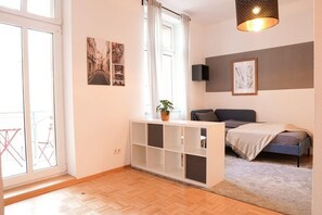 A modern bedroom with a bed, a console table with a plant, a large window with curtains, and artwork on the walls. - FullHouse | Halle | AB7 Medium Deluxe | Balkon (Halle (Saale))