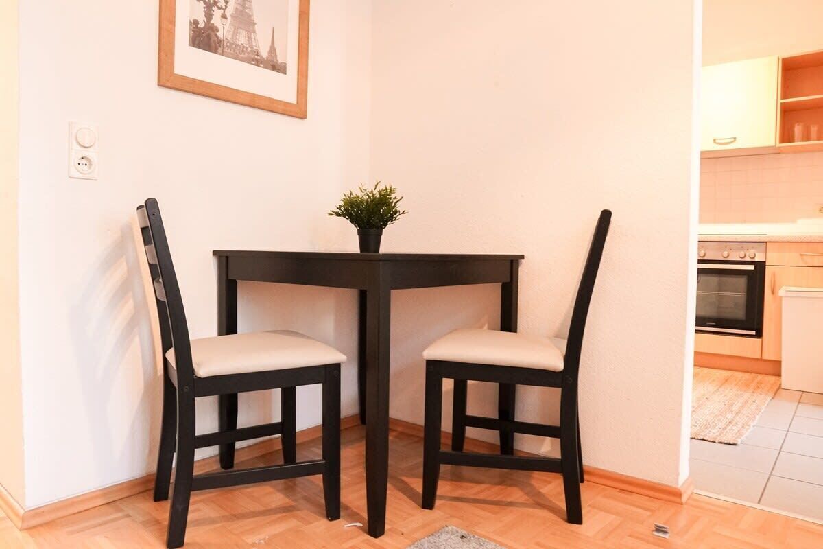Een gezellig eetgedeelte met een ronde tafel en twee stoelen, een potplant op de tafel en een ingelijste foto aan de muur.