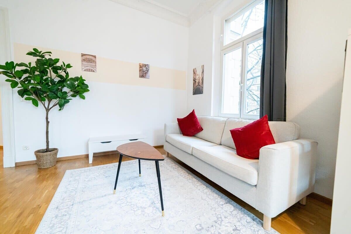 A modern living room with a white sofa, red cushions, a small wooden table, a potted plant, and a patterned rug.