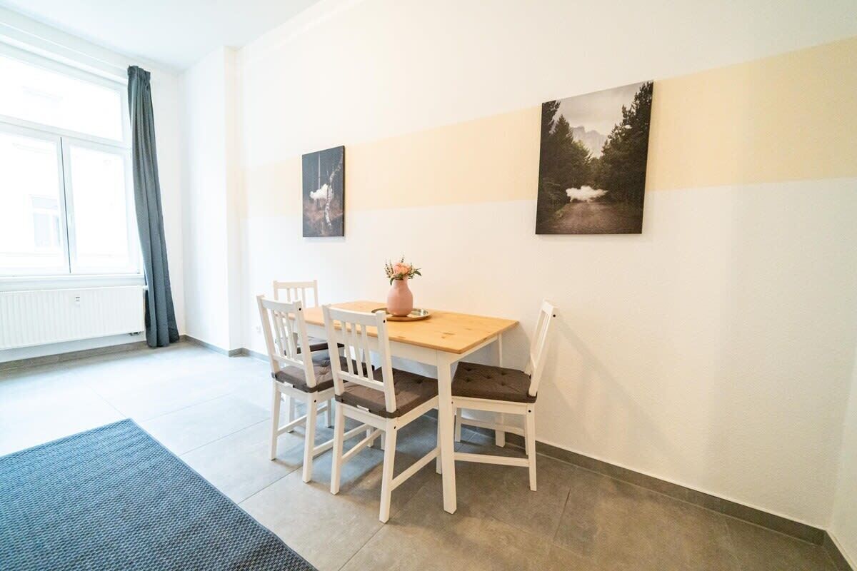 Une pièce avec une table en bois et quatre chaises blanches, un vase rempli de fleurs et deux tableaux encadrés accrochés au mur.