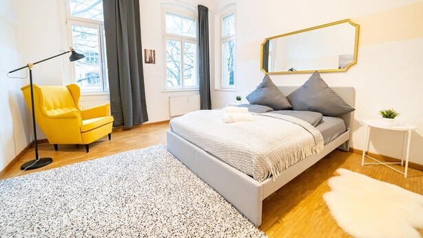 Un dormitorio con cama, una silla, una lámpara, un espejo y una mesita.