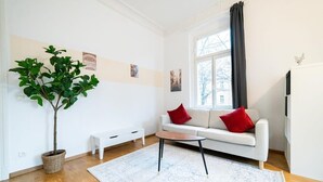 Una sala de estar moderna con un sofá blanco, cojines rojos, una mesita de centro, una planta en maceta y un amplio ventanal con cortinas.