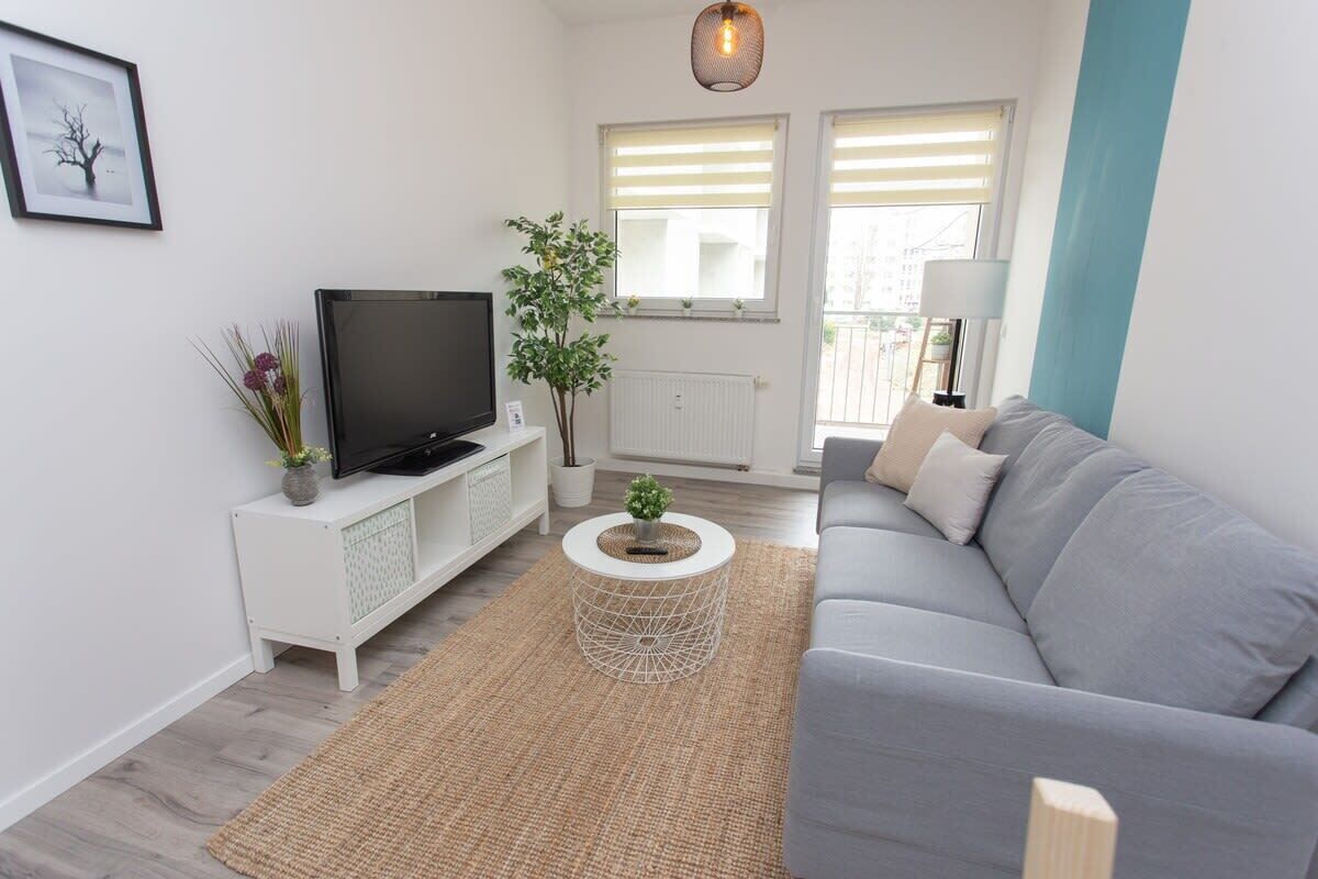 Een moderne woonkamer met een grijze bank, een televisie op een wit kastje, een ronde salontafel en een plant.