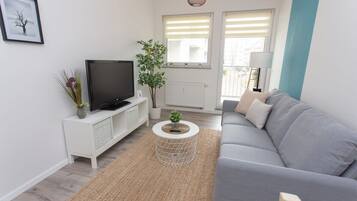 Een moderne woonkamer met een grijze bank, een televisie op een wit kastje, een ronde salontafel en een plant.