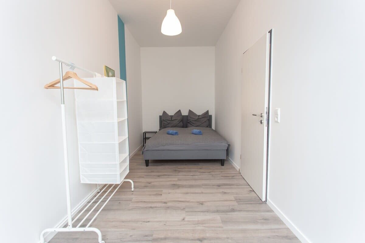 A narrow hallway leading to a bedroom with a bed, a white storage unit, and a hanging rack.