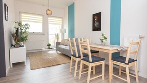 A modern living room with a wooden dining table, two chairs, a sofa, and a coffee table. There is a window with blinds, a lamp, and a plant on the windowsill. - FullHouse | Chemnitz | Rosenhof C8 | Zentrum (Chemnitz)
