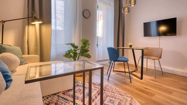 A modern living room with a glass coffee table, a sofa, a blue chair, a small table with a lamp, and a TV mounted on the wall. - FullHouse Premium Apt. H11 (Leipzig)