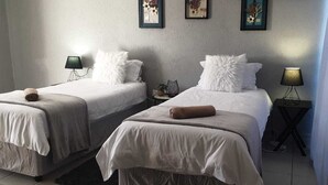 A hotel room with two beds, each with a white bedspread and a rolled towel. There are two bedside tables with lamps, and the walls are adorned with framed artwork. - Tsamatiku Guesthouse (Mangwazi)