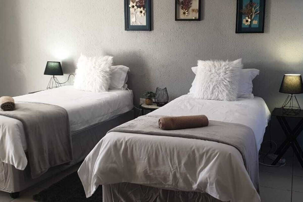 A hotel room with two beds, each with a white bedspread and a rolled towel. There are two bedside tables with lamps, and the walls are adorned with framed artwork.