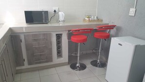 A kitchen with a microwave, kettle, and a refrigerator. There are two red bar stools and a white countertop. - Tsamatiku Guesthouse (Mangwazi)