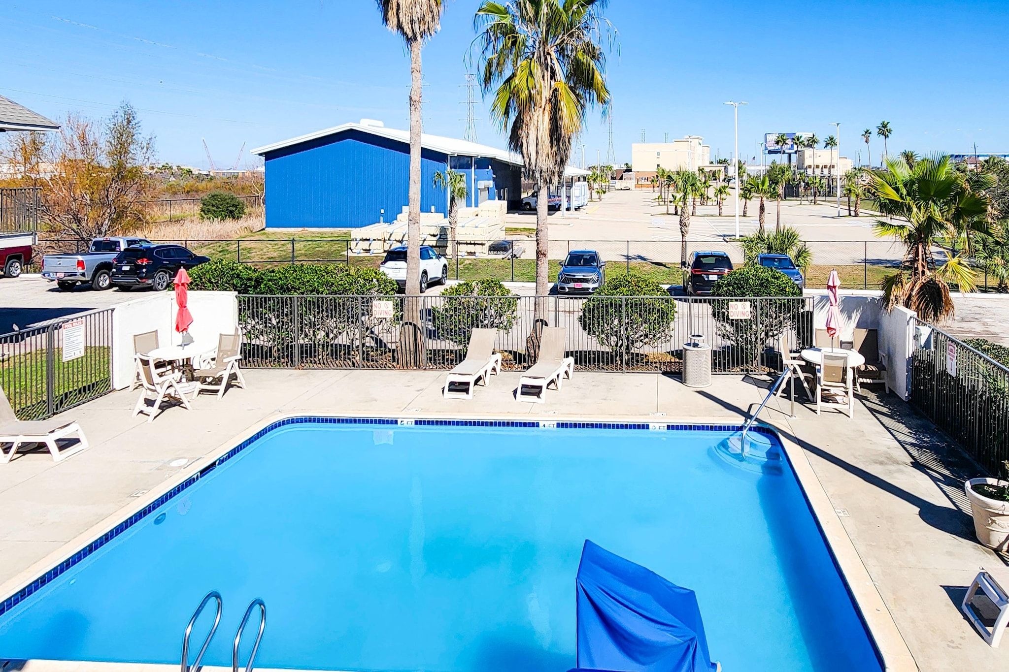 Piscina azul cristalina com espreguiçadeiras e guarda-sóis, cercada por palmeiras e um prédio azul ao fundo.