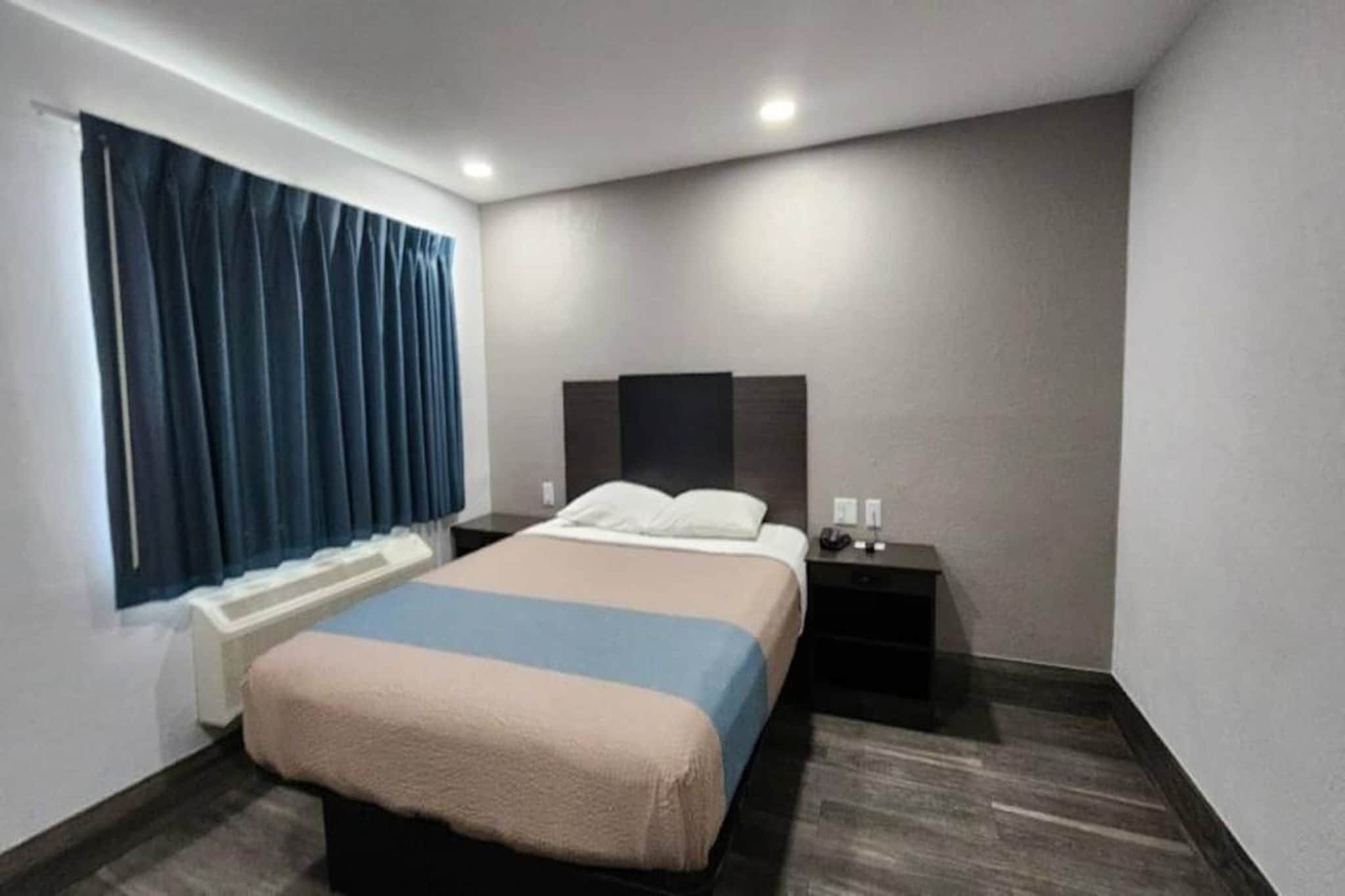 A hotel room with a bed, a nightstand, a window with curtains, and a wall-mounted heater.