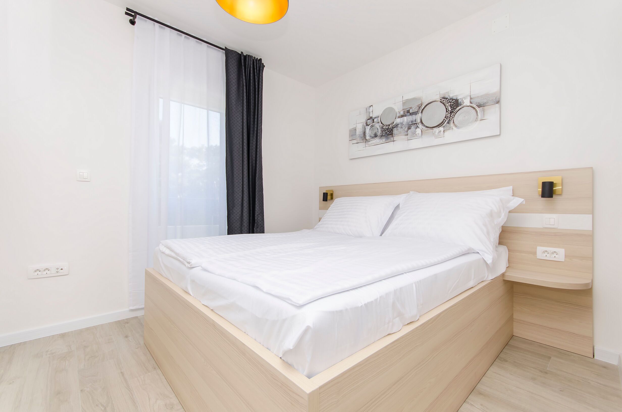 A neatly made bed with white linens in a modern room with light wooden flooring, a wall-mounted artwork, and a bedside table with a lamp.