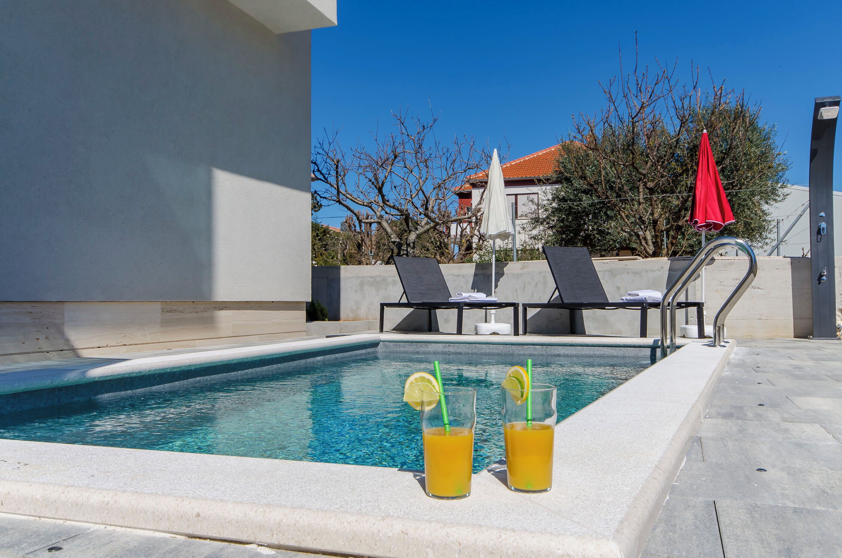A swimming pool with two glasses of orange juice, lounge chairs, and a red umbrella.