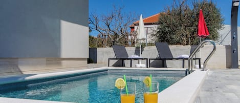 En swimmingpool med to glas appelsinjuice, liggestole og en rød parasol.
