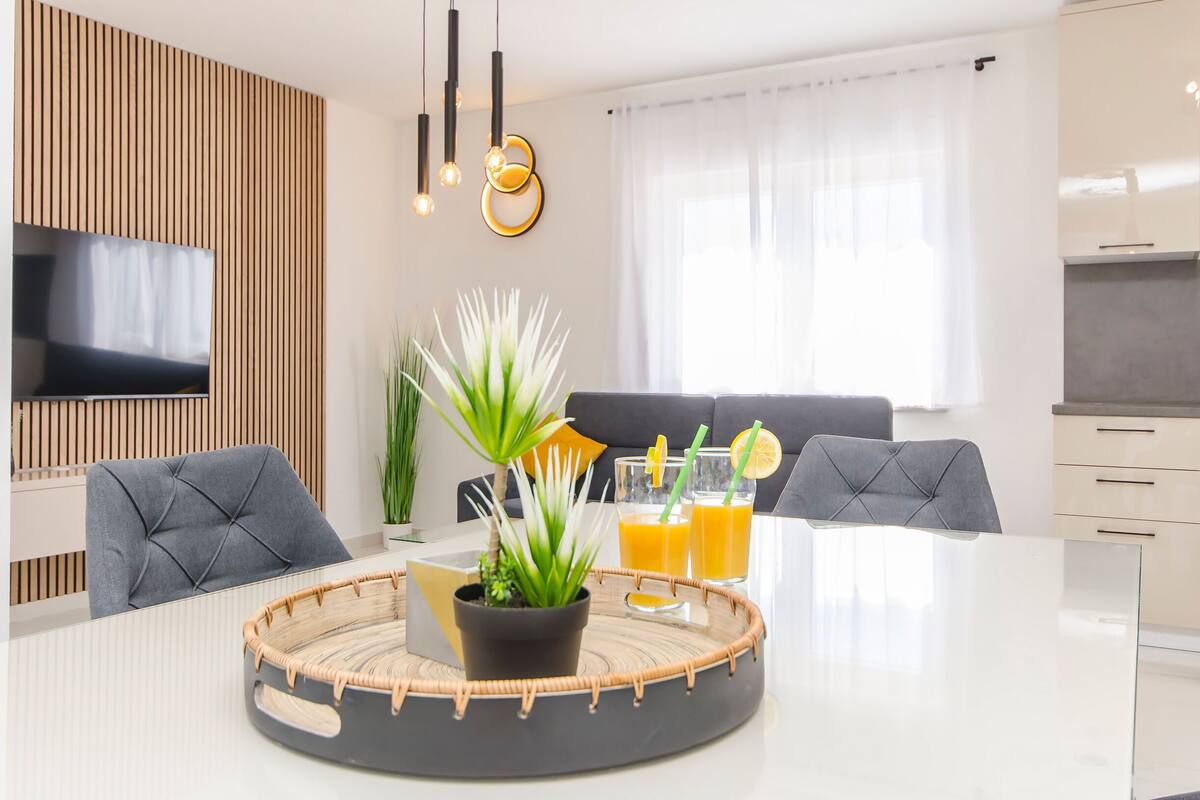 A modern living room with a round dining table, two grey upholstered chairs, a flat-screen TV mounted on a wooden panel, a potted plant, and a glass coffee table with a woven tray.