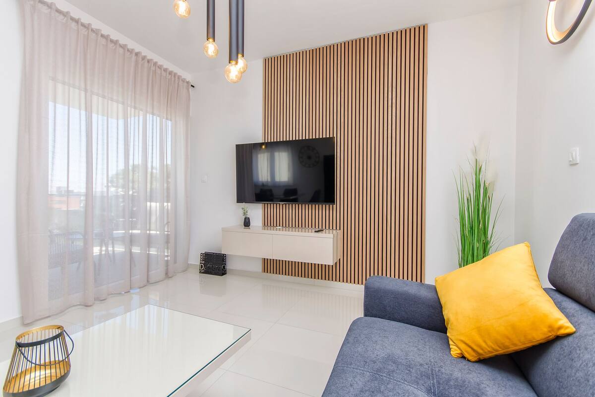A modern living room with a flat-screen TV, a sofa with a yellow pillow, a glass coffee table, and a plant.