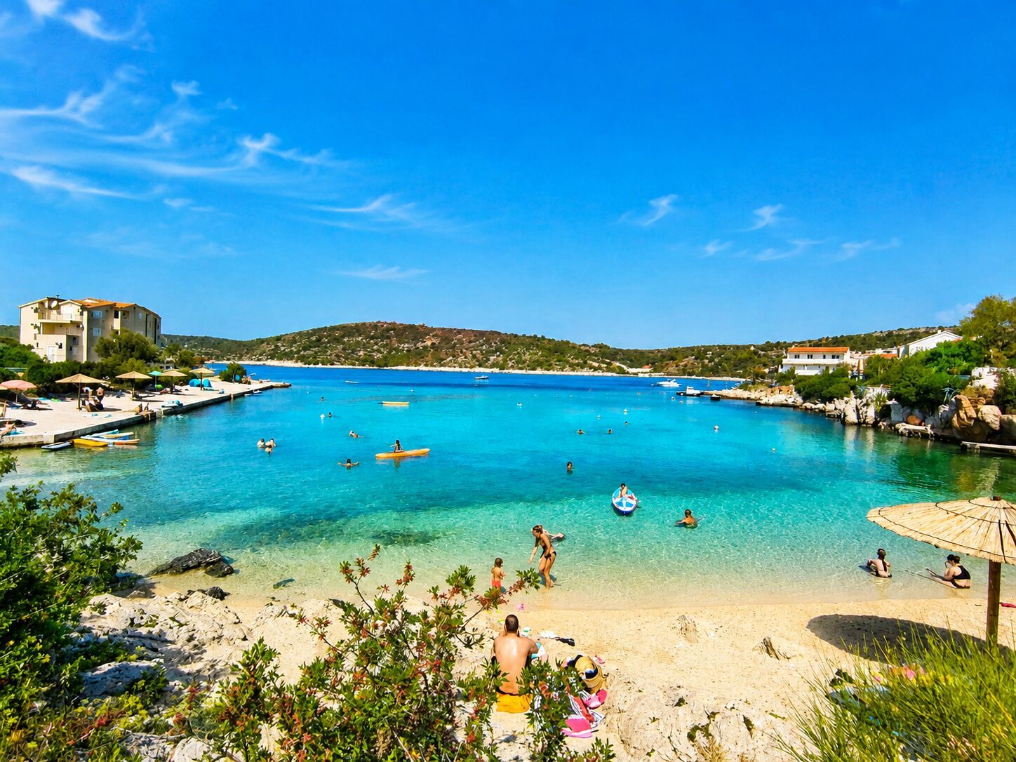 A coastal area with clear blue water, a sandy beach, and a few people swimming and sunbathing.