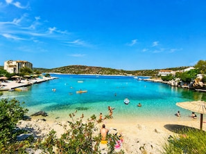 A coastal area with clear blue water, a sandy beach, and a few people swimming and sunbathing. - Apartments Neda (Rogoznica)