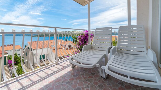 A balcony with two white lounge chairs, a tiled floor, and a view of buildings and the sea. - Apartments Teresa (Seget)