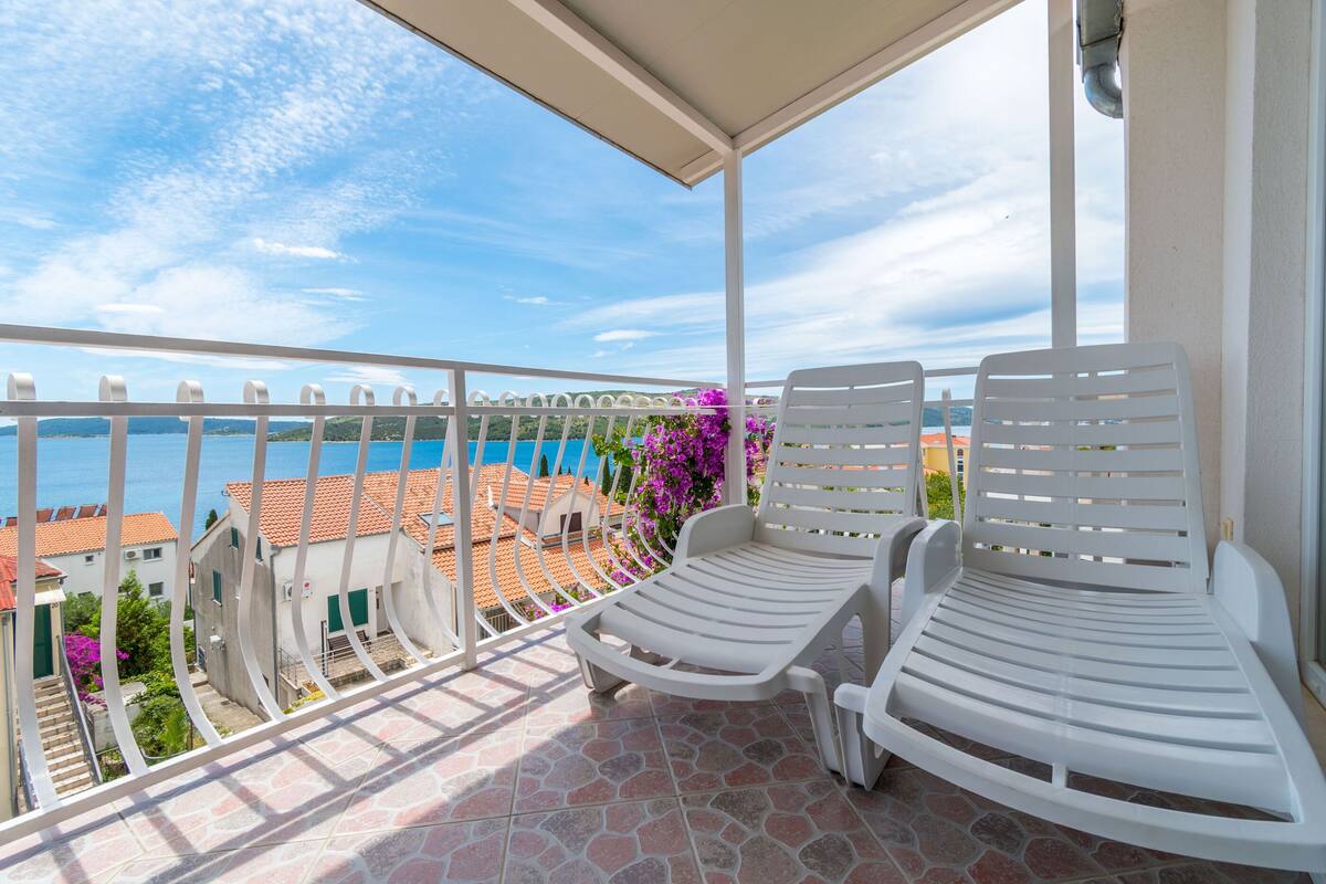 A balcony with two white lounge chairs, a tiled floor, and a view of buildings and the sea.