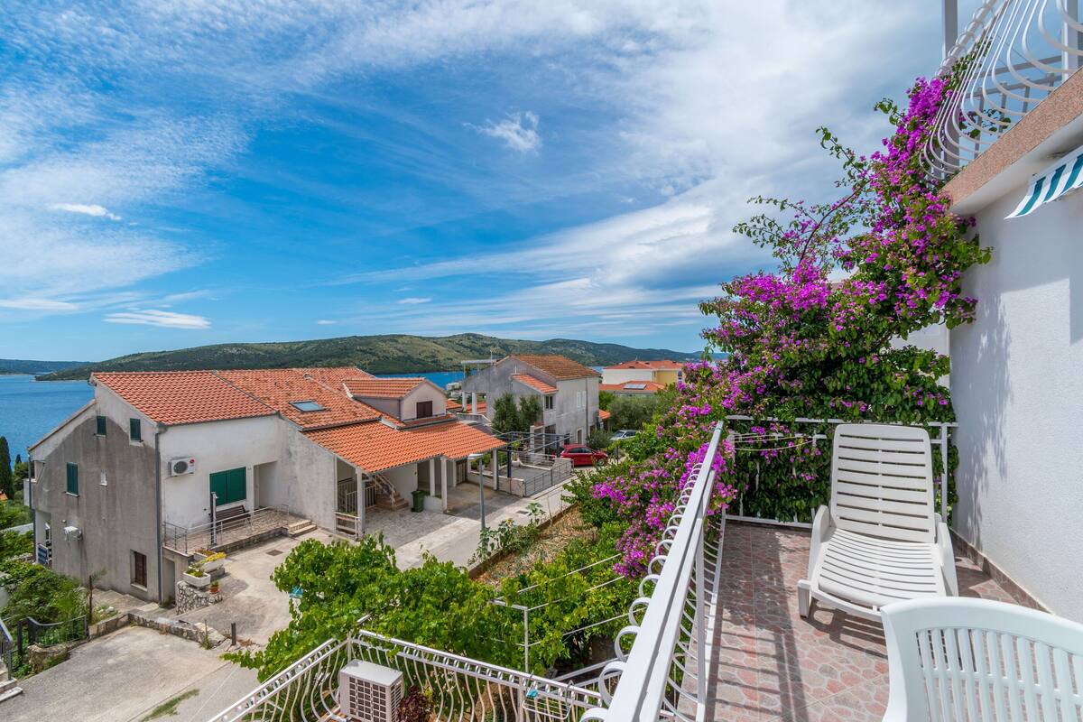 A balcony with a white chair, a view of a coastal village, and a building with pink flowering vines.