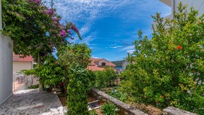 A stone-paved courtyard with a view of a red-tiled building, surrounded by lush greenery and flowering plants. - Apartments Teresa (Seget)
