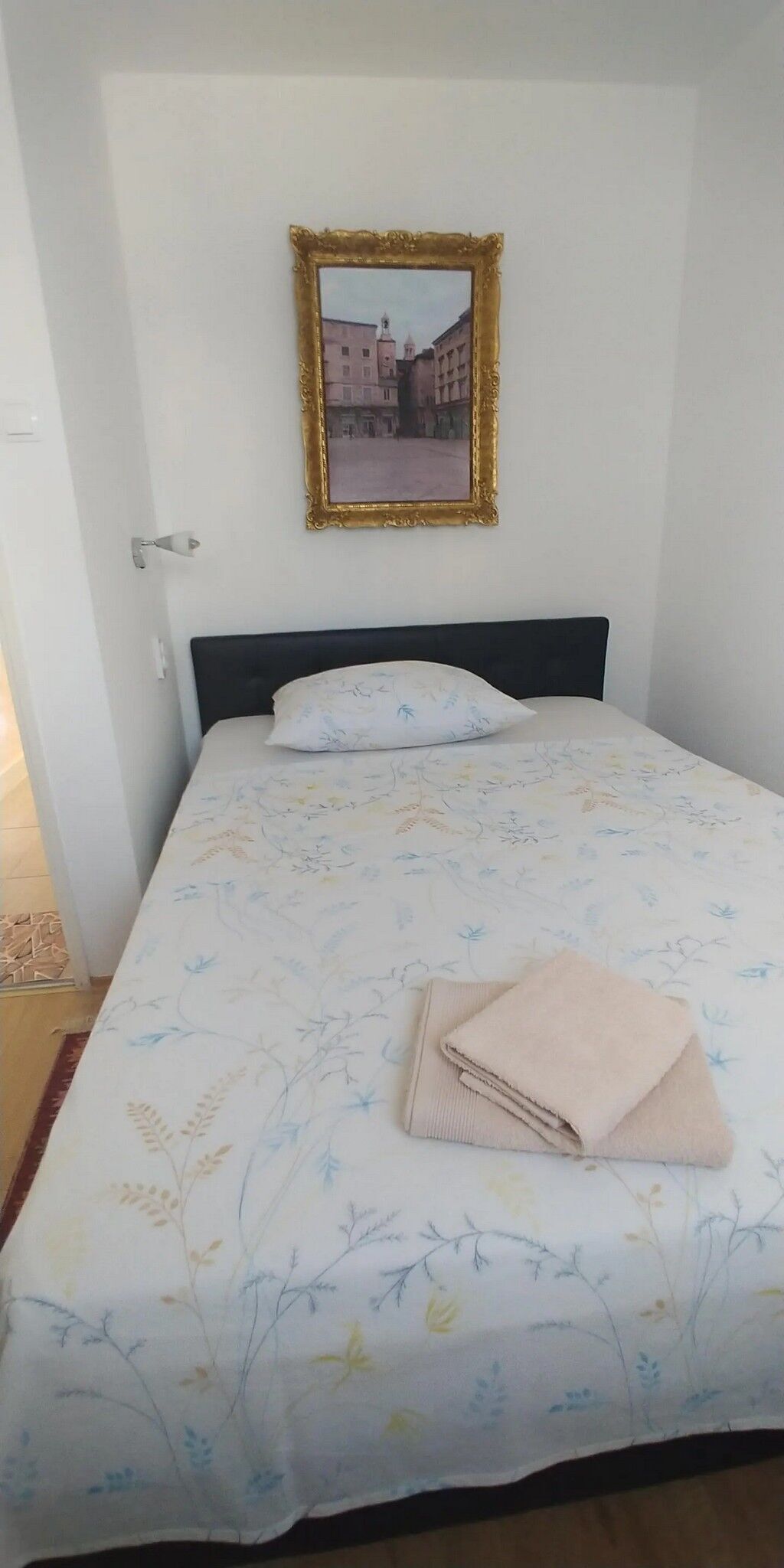 Ein ordentlich bezogenes Bett mit einer blumigen Tagesdecke, ein gefaltetes Handtuch und ein gerahmtes Bild eines Gebäudes an der Wand.