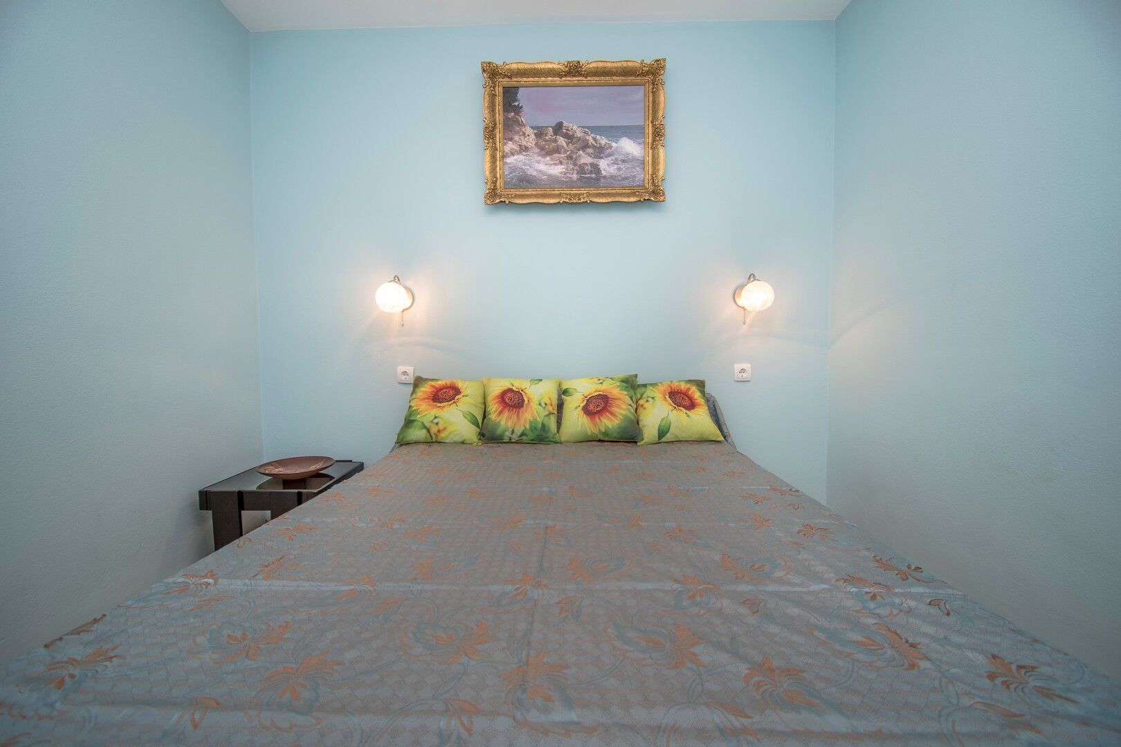 A bedroom with a bed, a nightstand, a wall-mounted picture frame, and two wall sconces.
