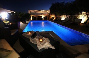Zona de piscina nocturna con sillones de descanso y una mesa con velas y un teléfono.