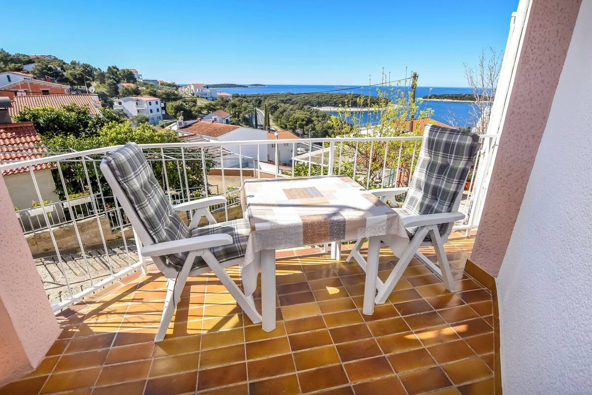 A balcony with two white chairs and a table, overlooking a coastal town and the sea.