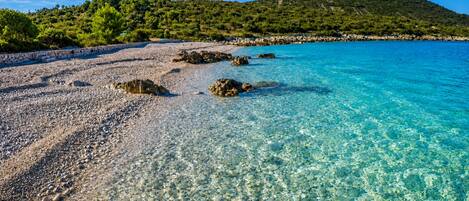A clear beach with pebbles and rocks, a calm sea, and a green hill in the background.