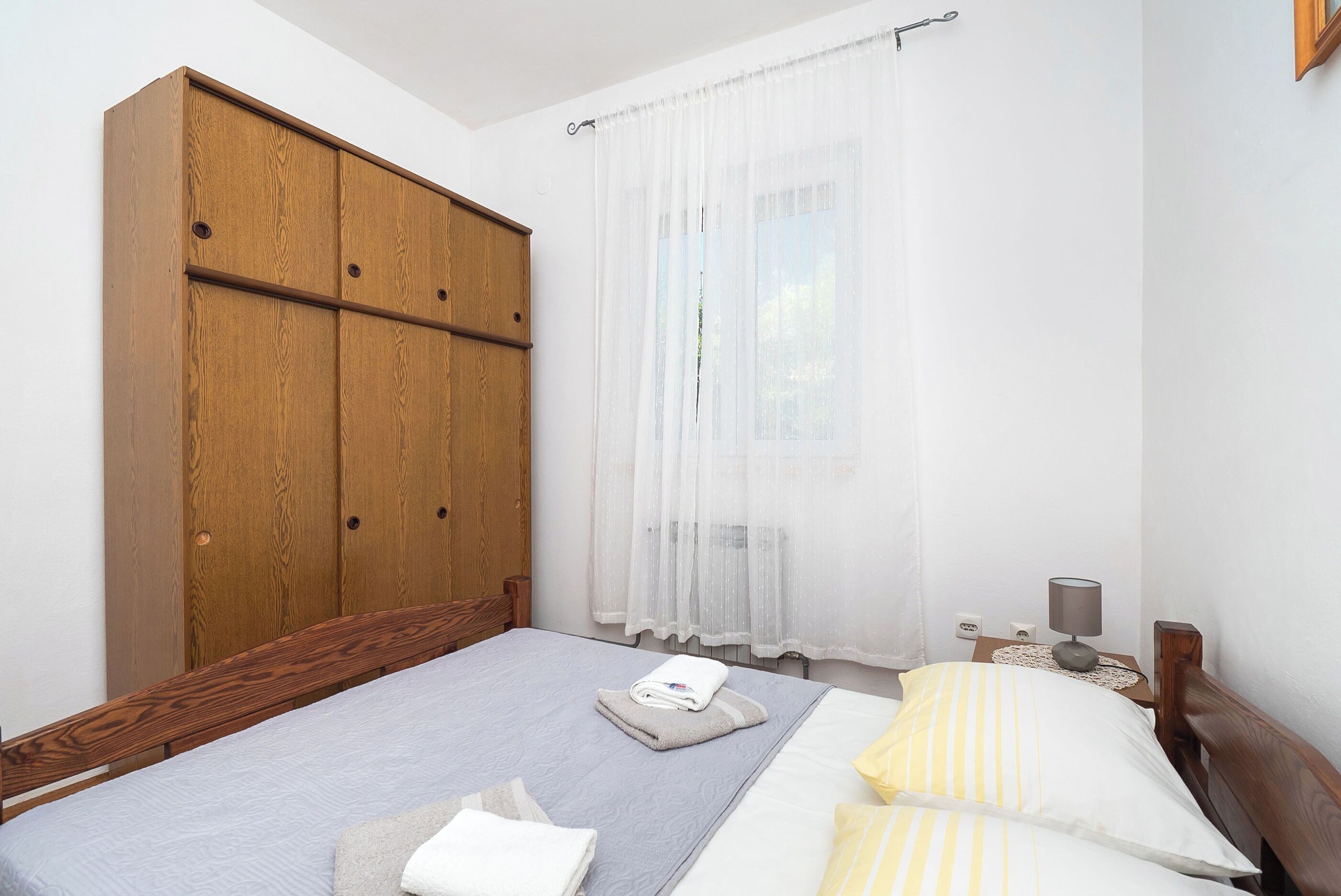 Une chambre à coucher avec une armoire en bois, un lit recouvert d’une couette à rayures grises et jaunes, et une table de chevet avec une lampe.