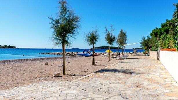 Een pad van stenen tegels langs het strand, met bomen, strandstoelen en parasols.