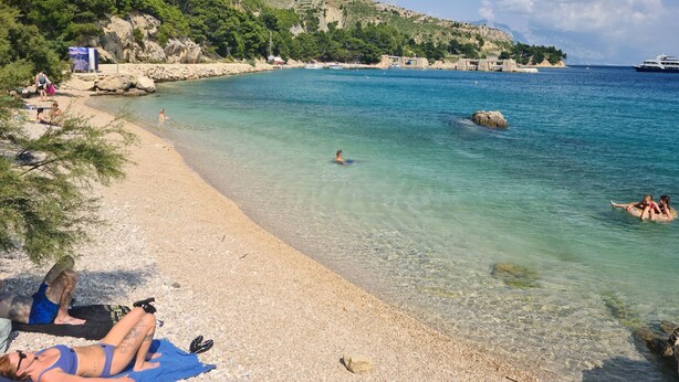 A beach with clear water, rocky shore, and a boat in the distance. - House Pine Tree (Omis)