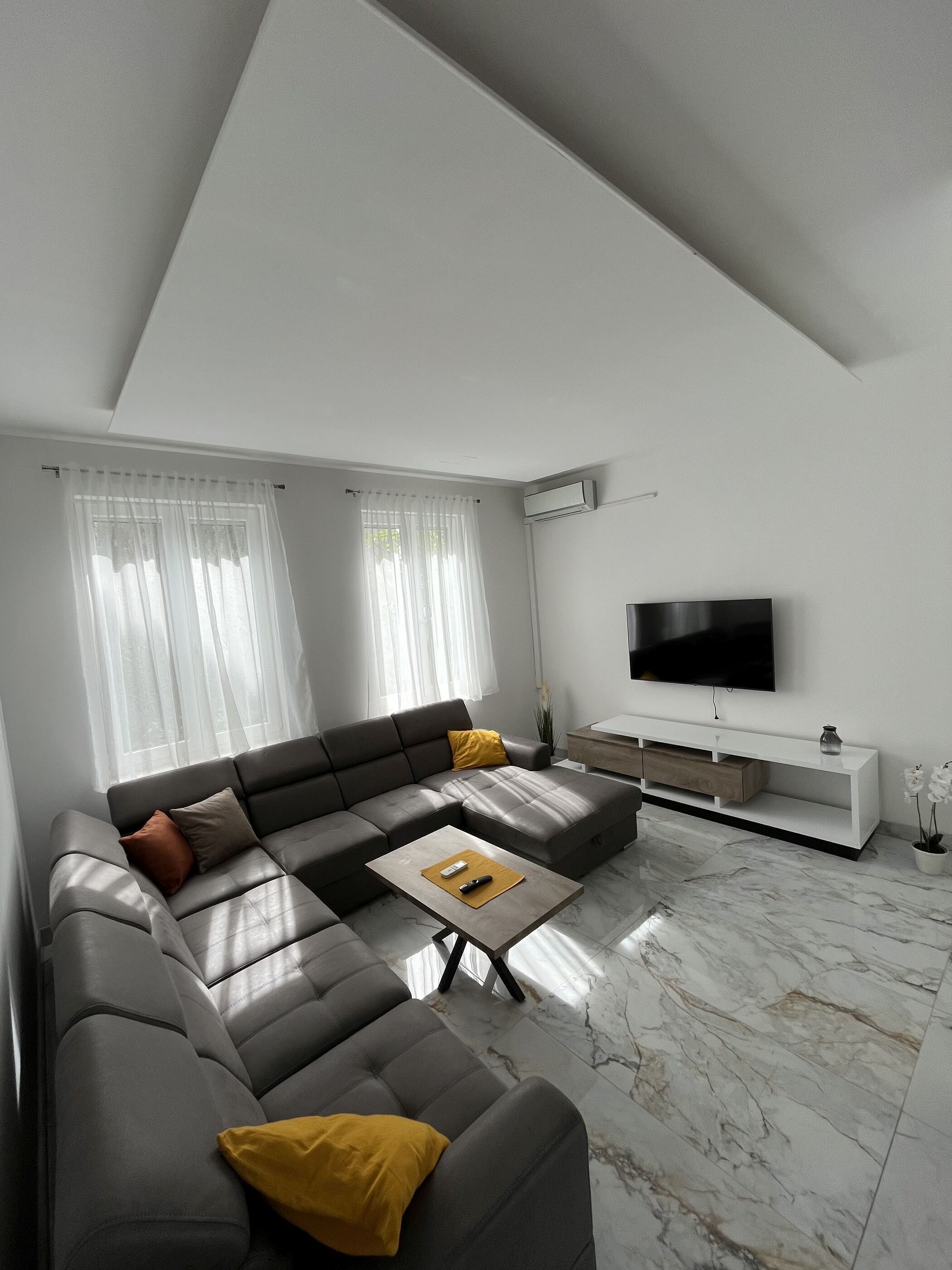 Un salon moderne avec un canapé d’angle gris, un téléviseur à écran plat, une table basse en bois et un sol en marbre.