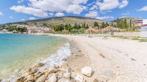 Una playa con agua turquesa cristalina, costa rocosa y un paisaje montañoso al fondo con edificios y árboles.