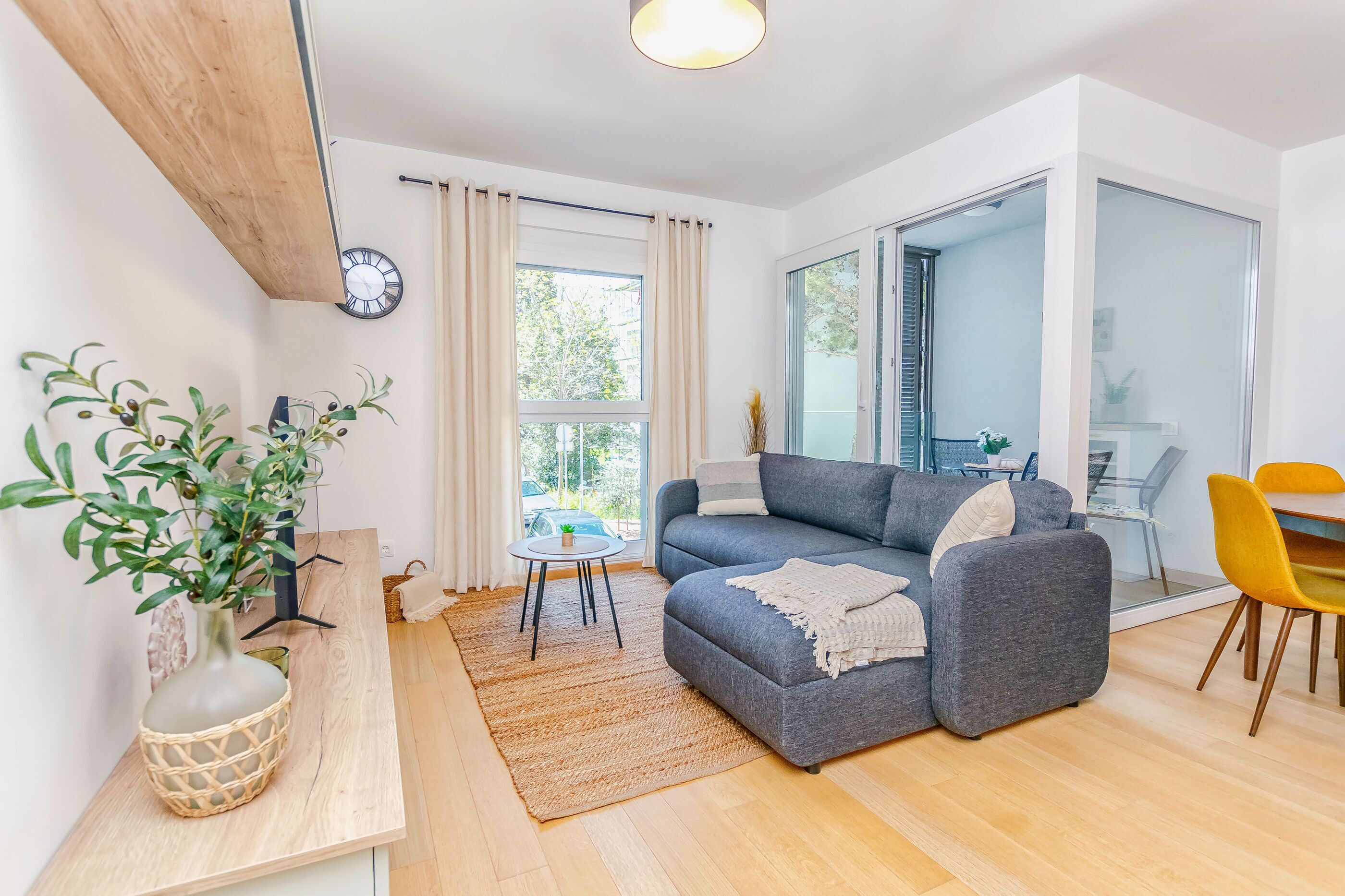 A modern living room with a grey sofa, a small round table, and a yellow chair. There is a wooden floor, a large window with curtains, and a plant in a woven basket.