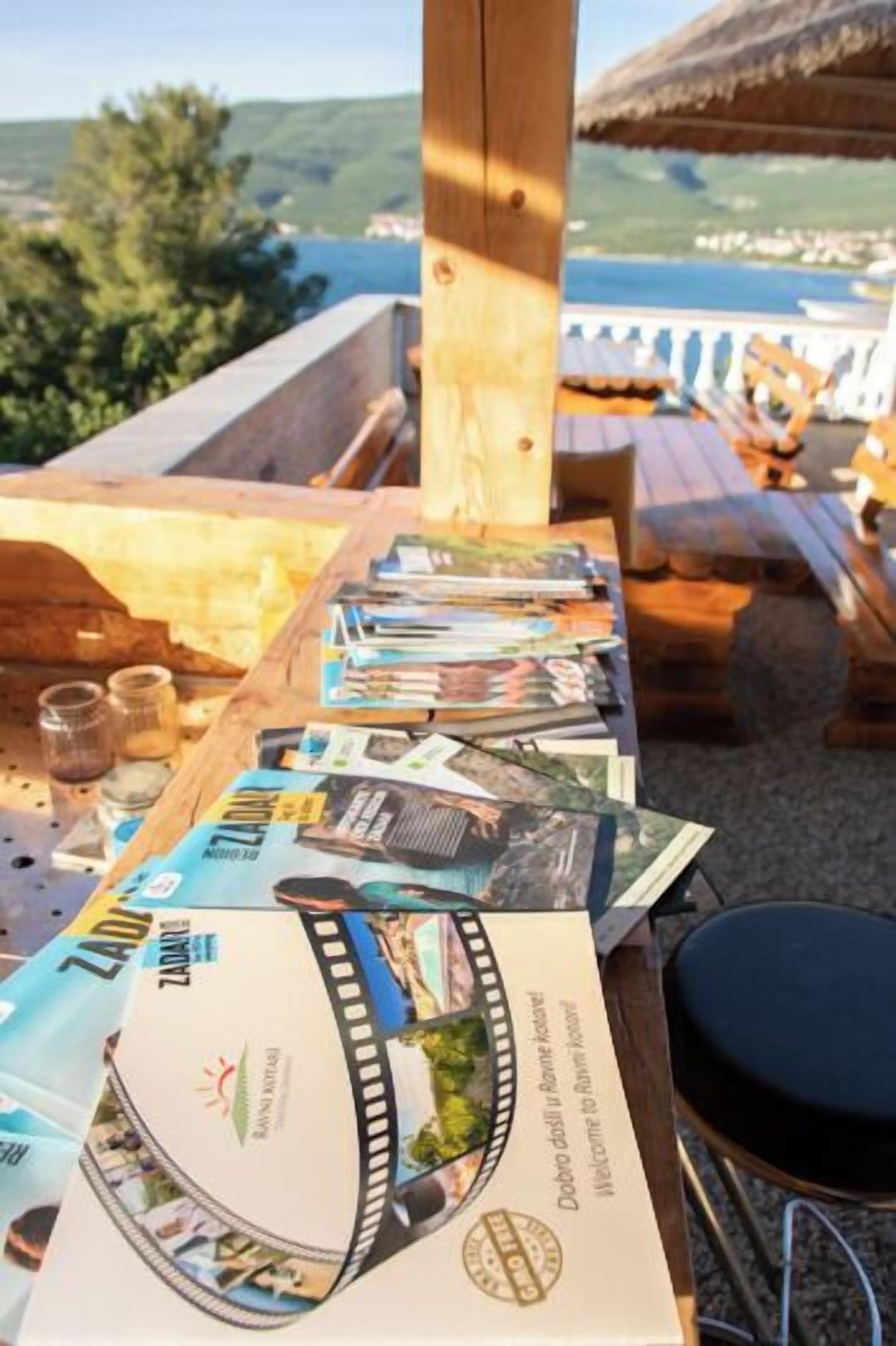 A wooden table with magazines and a ZADAR brochure, set outdoors with a view of the sea and hills.