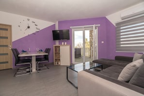 A modern living room with a sofa, a dining table with chairs, a TV mounted on the wall, and a sliding glass door leading to an outdoor area. - Apartments Kristina (Benkovac)