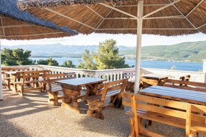 Een terras met houten tafels en banken, een rieten parasol en uitzicht op een meer en bergen.