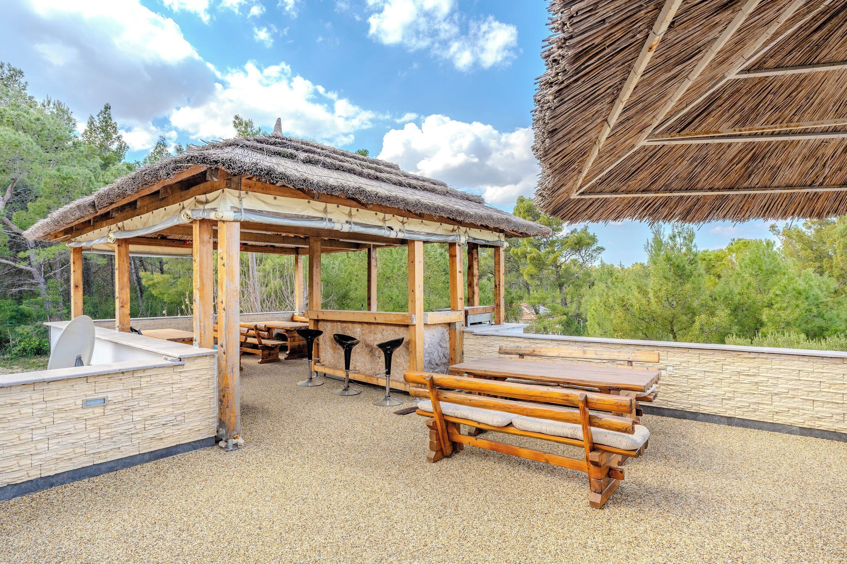 A wooden gazebo with a thatched roof, surrounded by a stone wall and equipped with picnic tables and benches.